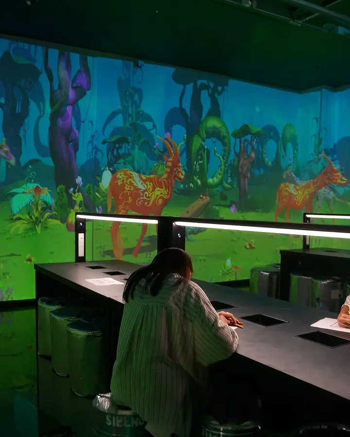 A visitor colours a drawing at the tables under the projected wall.
